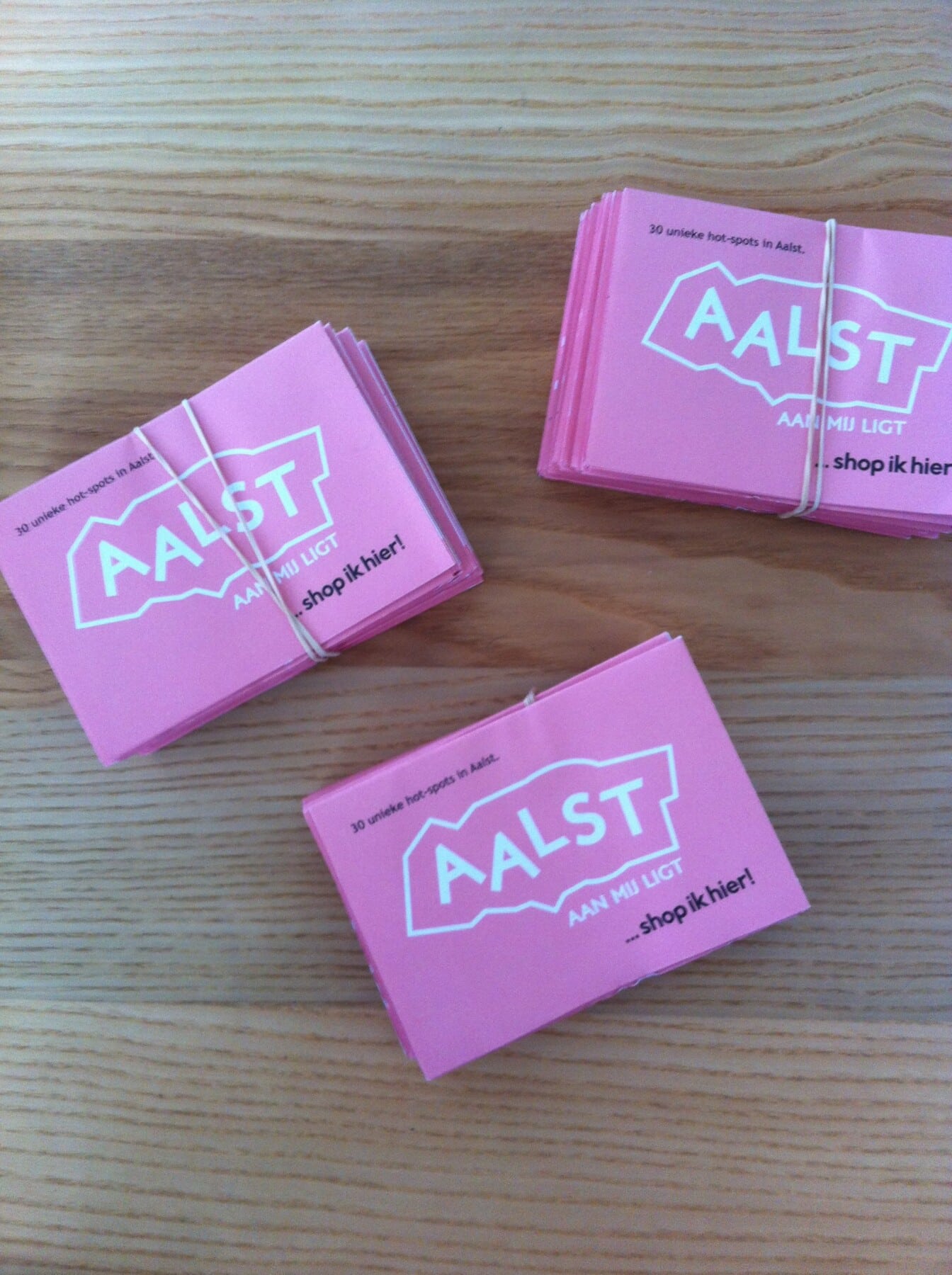 Drie roze stadskaarten van Aalst met "AALST AAN MIJ LIGT" logo liggen op houten tafel