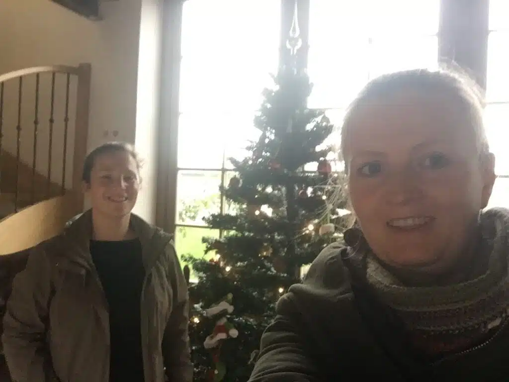 Twee vrouwen poseren lachend voor een versierde kerstboom in een gezellige woonkamer tijdens winterverblijf