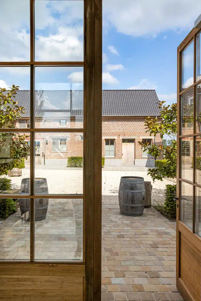 Uitzicht door openslaande deuren op binnenplaats van gerenoveerde hoeve Eyndevelde met houten vaten en bakstenen gebouwen