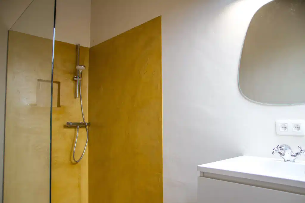 Moderne badkamer met geel accent muur, glazen douchewand, ronde spiegel en witte wastafel in Eyndevelde vakantiewoning