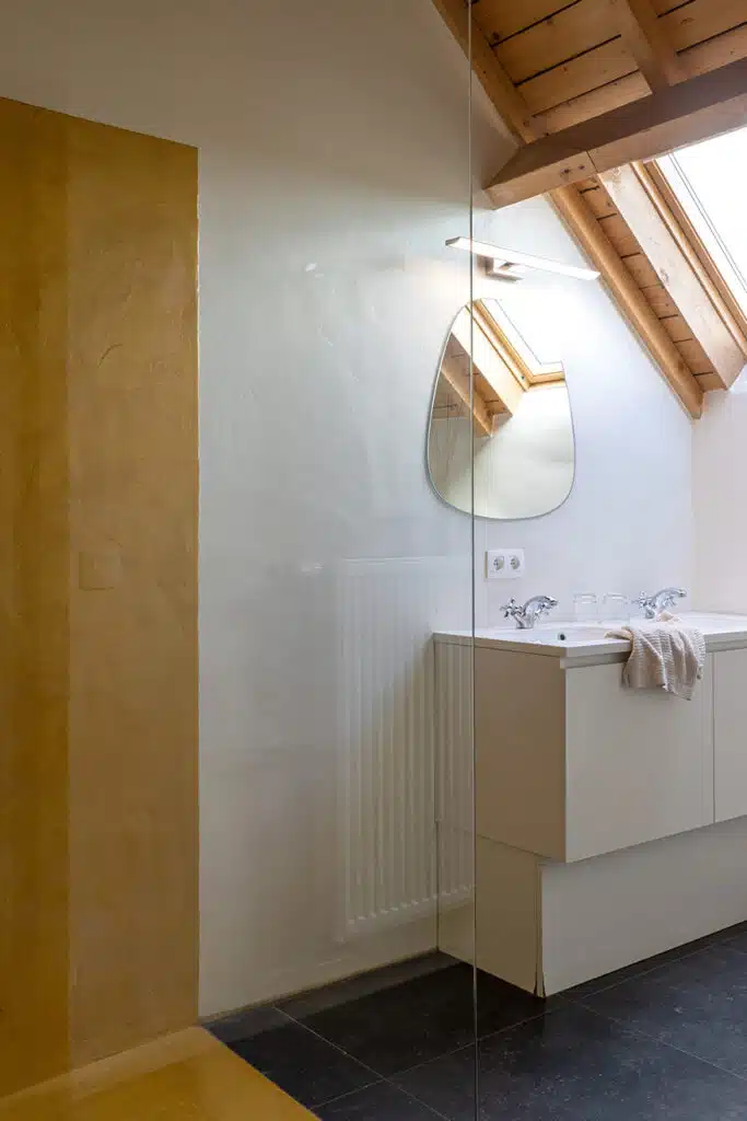 Luxueuze badkamer met houten balken, glazen douchewand, wit bad en ronde spiegel in Eyndevelde vakantiewoning