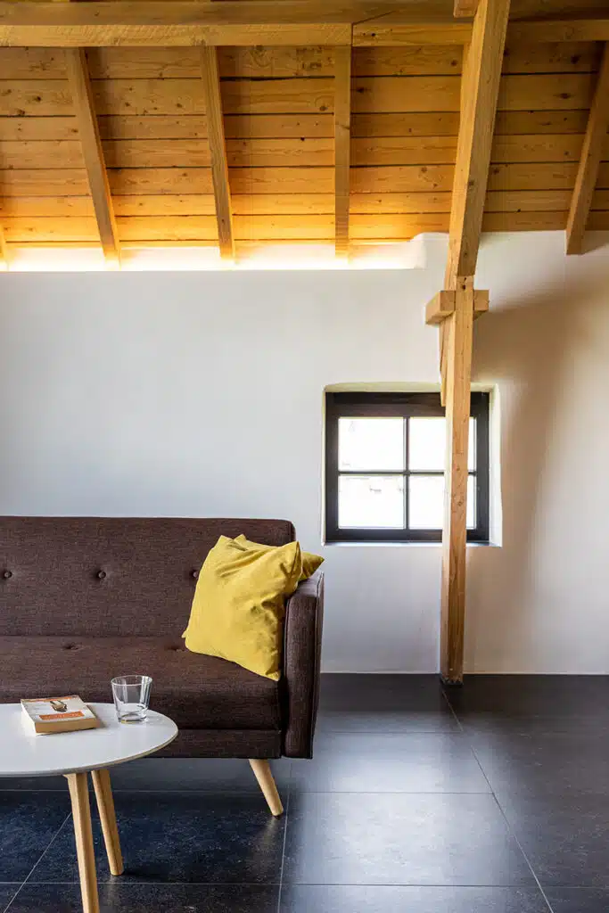 Moderne woonkamer in vakantiewoning Eyndevelde met houten balken, bruine bank, gele kussens en wit rond tafeltje