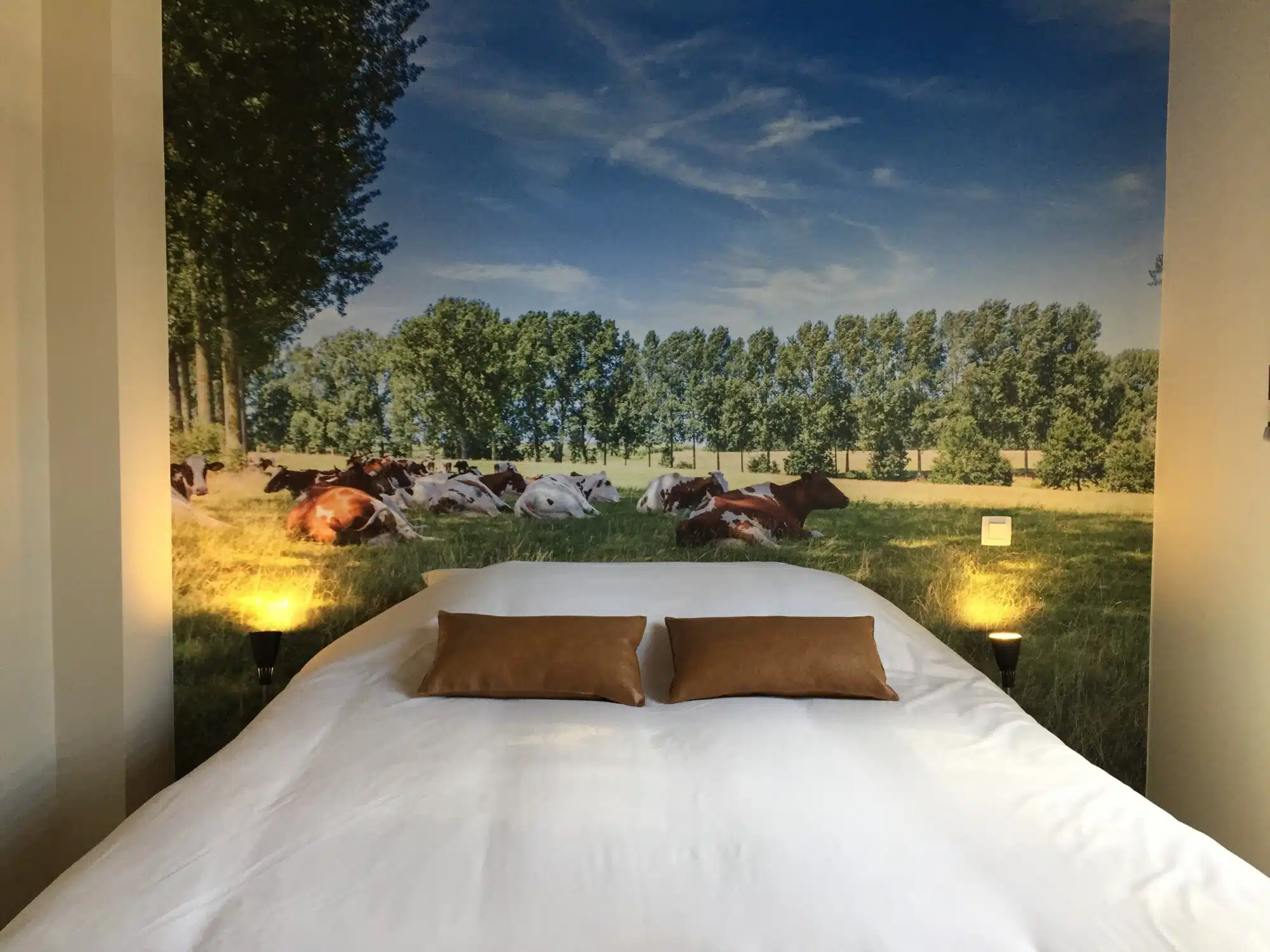 Slaapkamer met wit bed en kussens, muurafbeelding van koeien in weiland bij Eyndevelde vakantiewoning