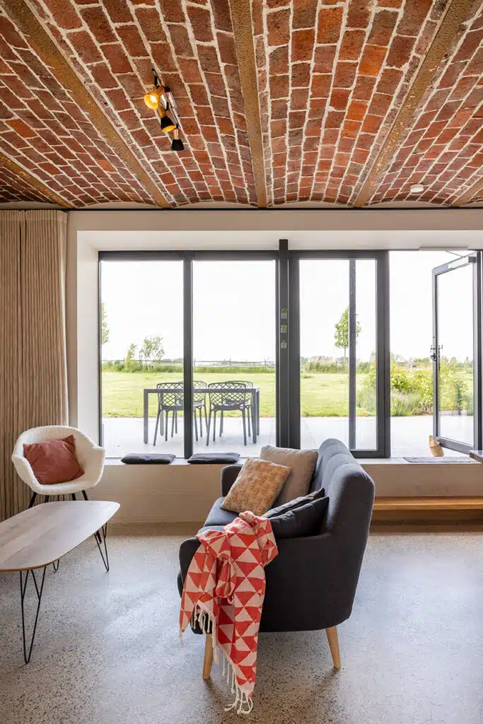 Moderne woonkamer met bakstenen gewelfplafond en uitzicht op groene tuin in Eyndevelde vakantiewoning