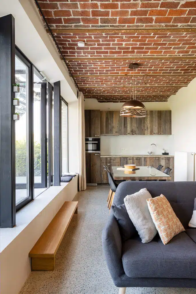 Moderne woonkamer met bakstenen plafond, houten keuken en grijze bank in Eyndevelde vakantiewoning