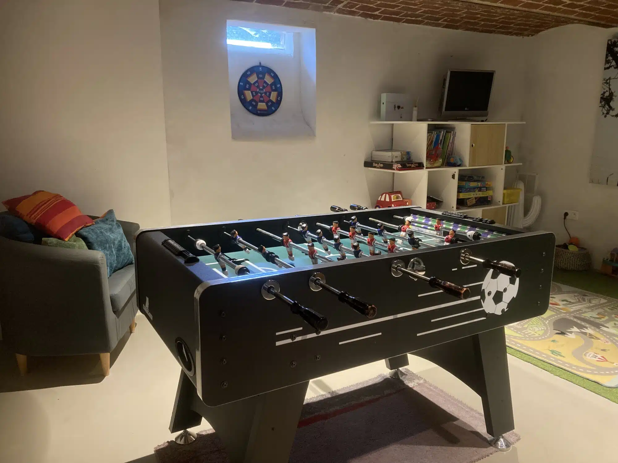 Speelkamer met zwarte tafelvoetbaltafel, grijze bank, speelgoed en projectie aan de muur in vakantiewoning