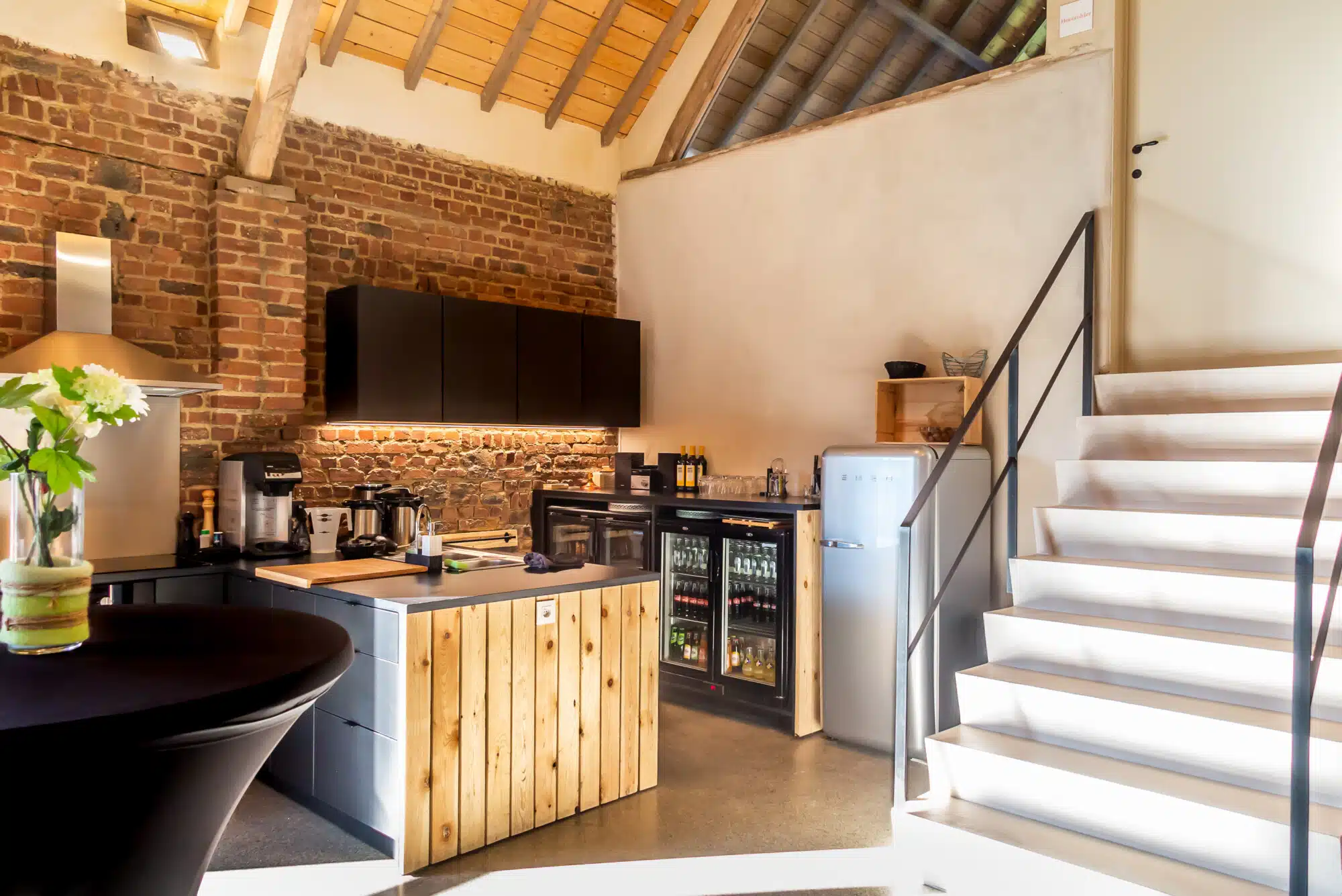 Moderne groepskeuken met bakstenen muur, houten keukeneiland en trap in Eyndevelde vakantiewoning