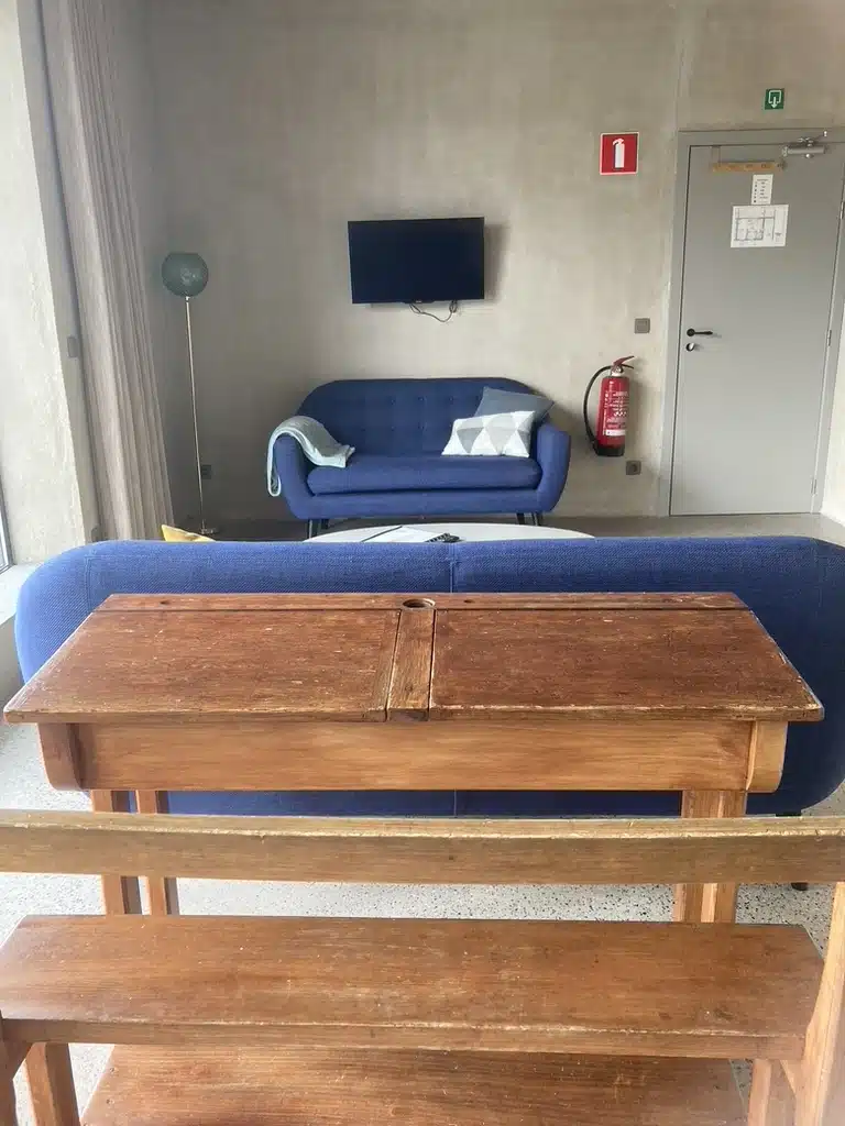 Gezellige gemeenschappelijke ruimte bij Eyndevelde met blauwe sofa's, houten tafel, muurTV en brandblusser aan de muur