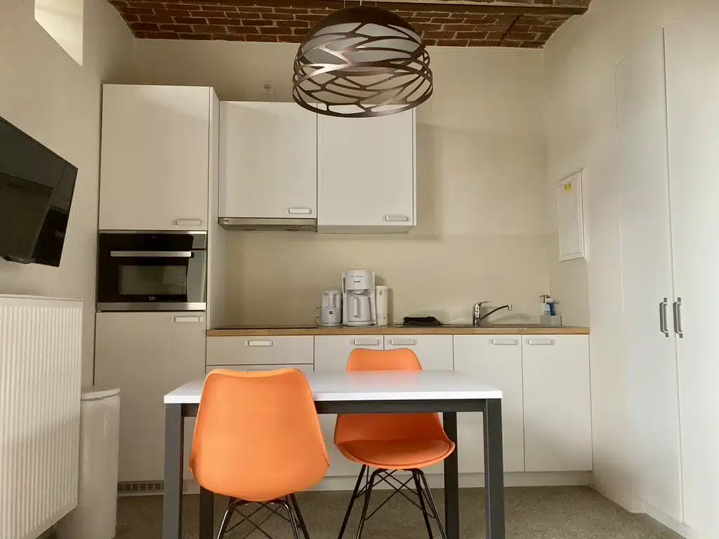 Moderne keuken met witte kasten, houten aanrecht, koffiemachine, oven en witte eettafel met oranje stoelen bij Eyndevelde