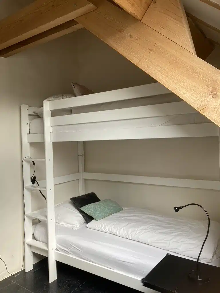 Witte stapelbed met ladder onder houten dakbalken in een slaapkamer van Eyndevelde, met witte beddengoed en kleurrijke kussen
