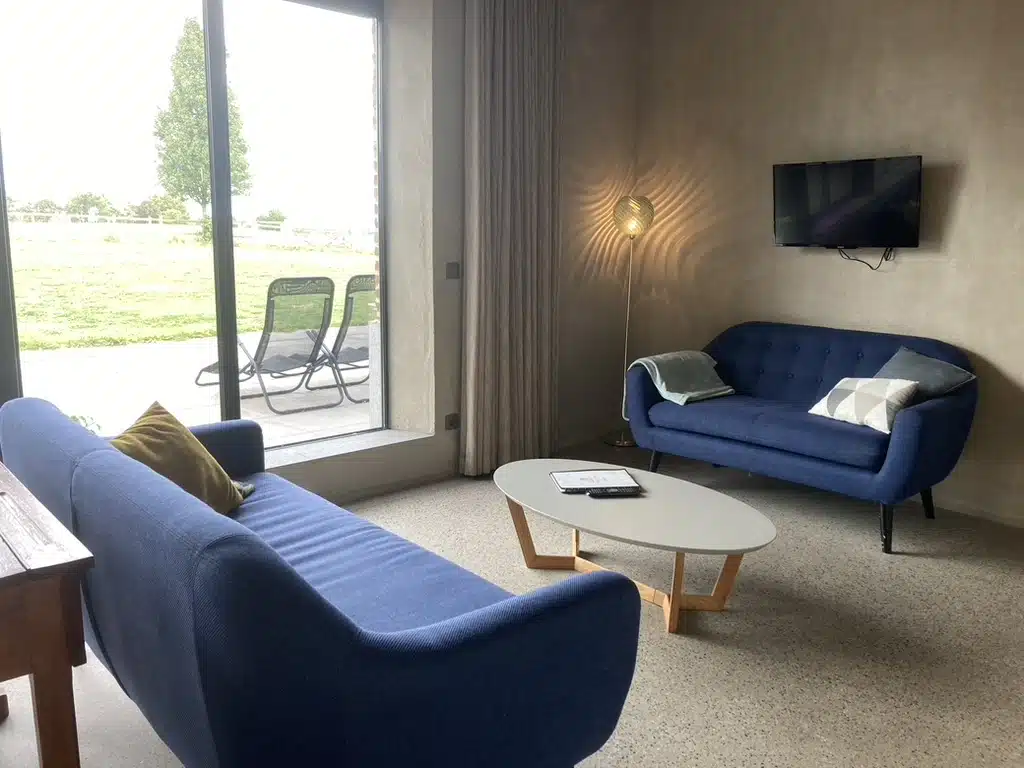 Gezellige woonkamer bij Eyndevelde met blauwe sofa's, salontafel, wandtv en grote schuifpui met zicht op het groene landschap