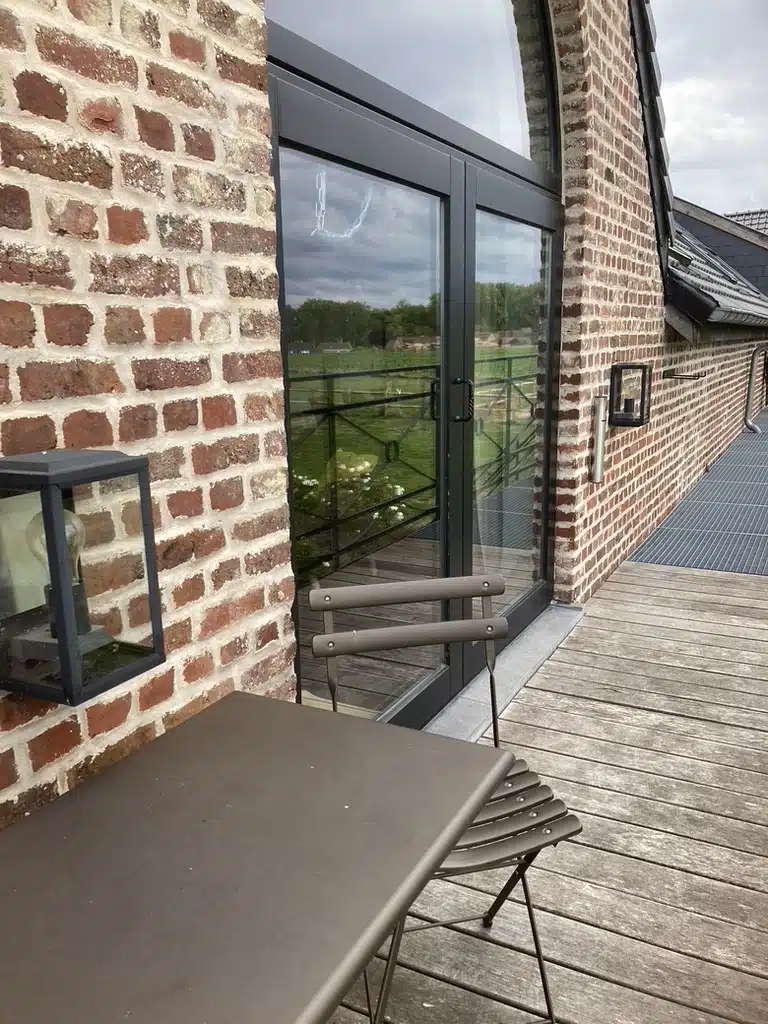 Houten terras bij Eyndevelde met bistrostoelen, glazen schuifdeur en rustieke bakstenen muur met zicht op groene velden
