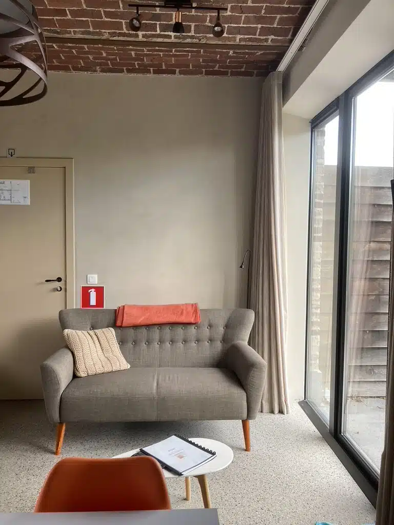 Gezellige zithoek bij Eyndevelde met grijze tweezitbank, kussens, grote ramen met gordijnen en exposed brickwork plafond