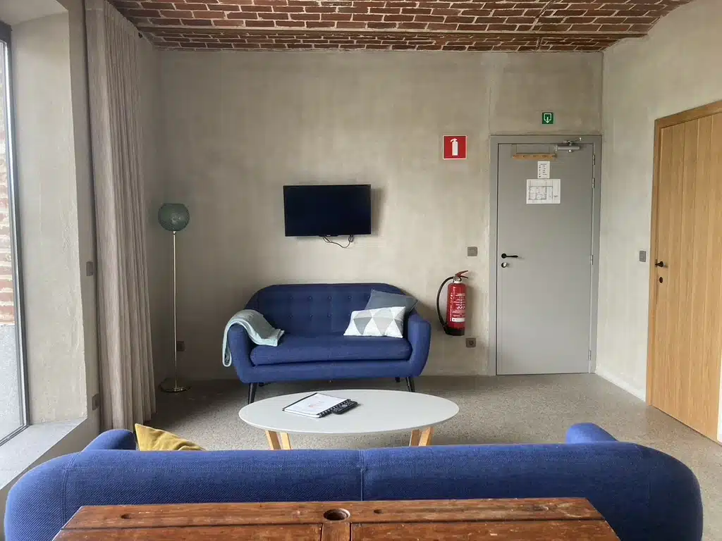 Gezellige woonruimte bij Eyndevelde met blauwe bankstellen, wandtelevisie, salontafel en bakstenen plafond — ideaal voor onts